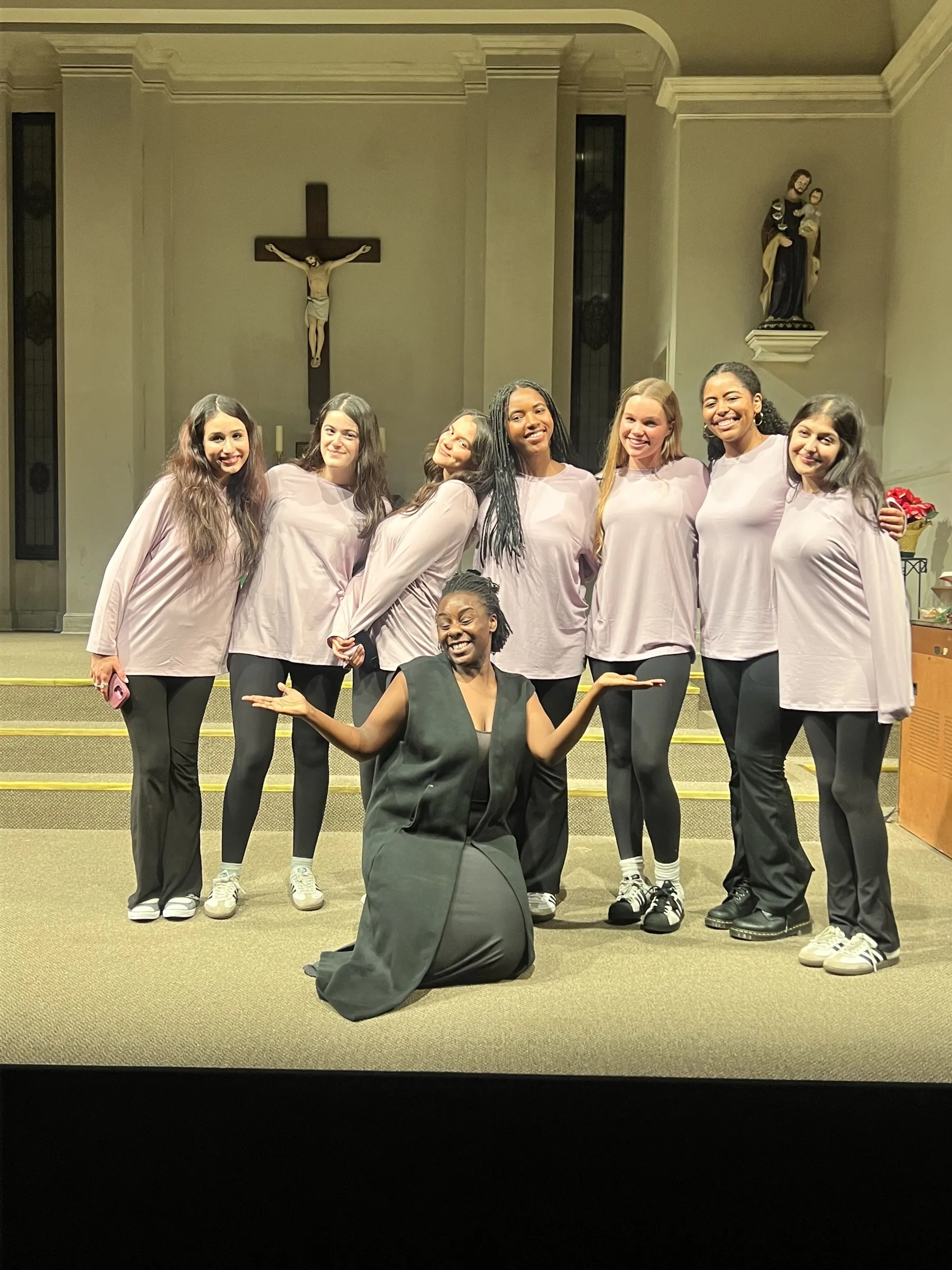 Abigail kneeling at the front of a chapel stage with her arms open, seven students in matching lilac tops standing behind her